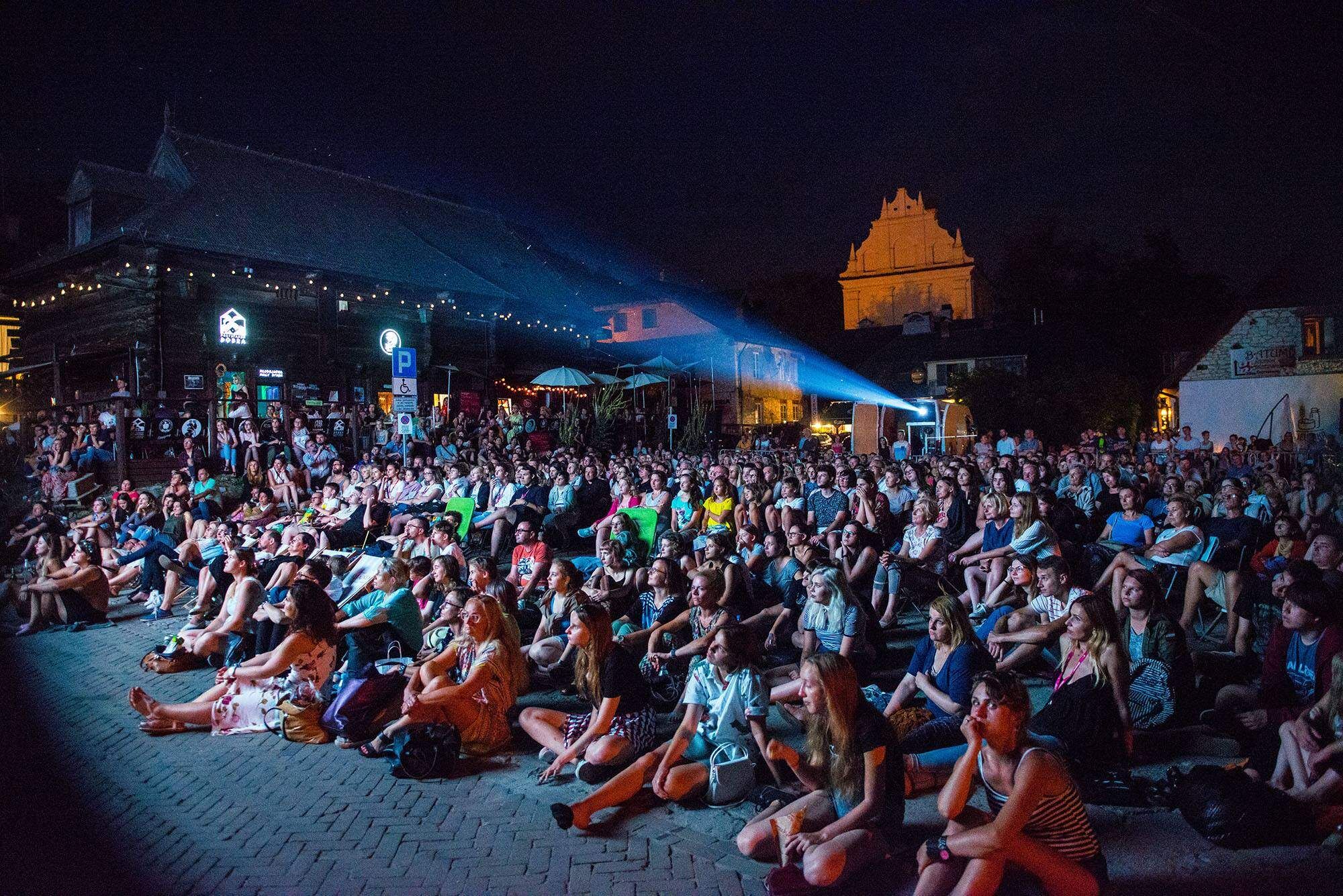 Duża grupa ludzi siedzi na krzesłach i ziemi pod gołym niebem, oglądając projekcję na ekranie. W tle widać oświetlone budynki i ciemne niebo. Atmosfera sugeruje plenerowe wydarzenie filmowe.