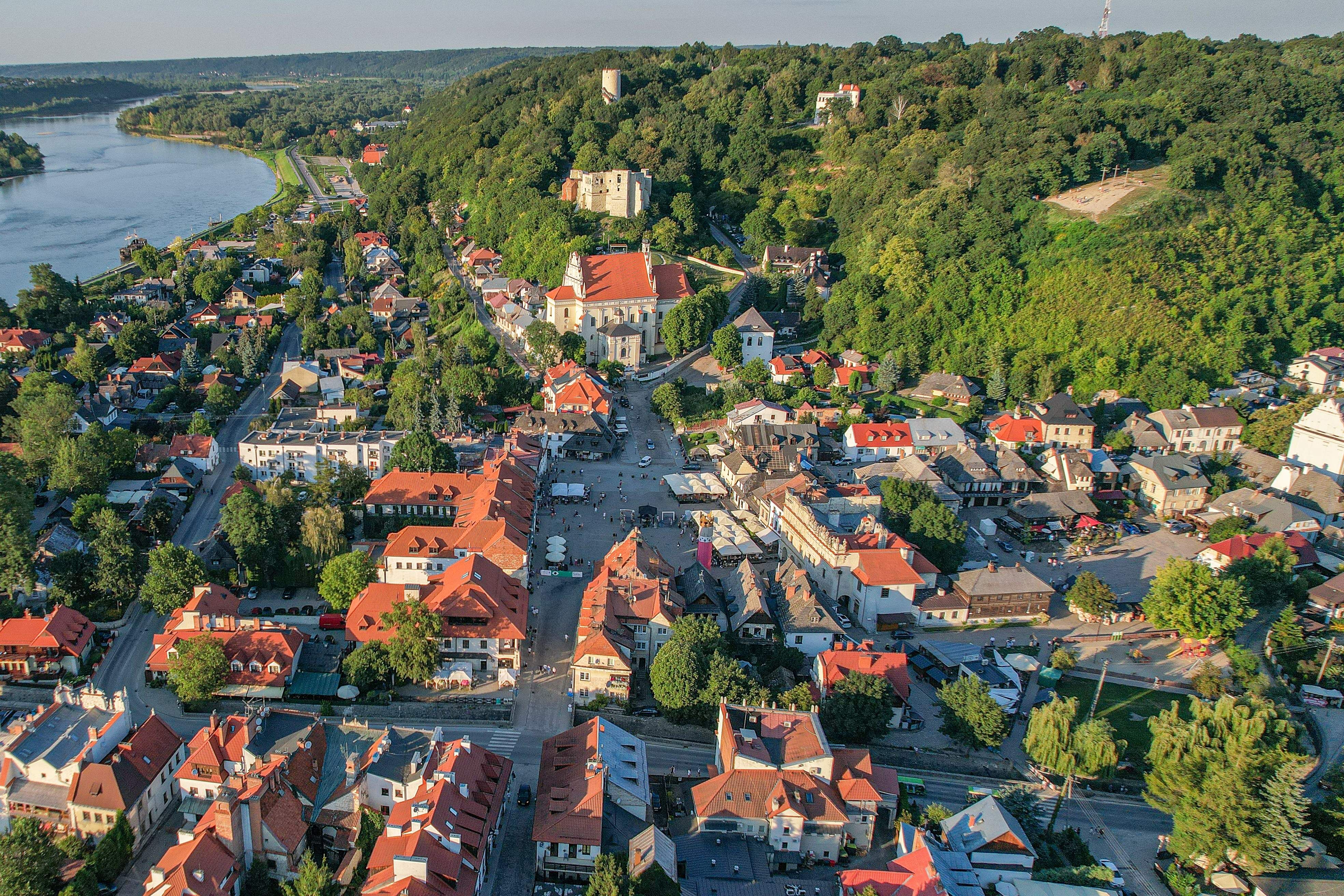 Widok z lotu ptaka na malownicze miasteczko z czerwonymi dachami, położone nad rzeką. W centrum widać urokliwy rynek z kościołem, otoczony zielonymi wzgórzami i zabytkowym zamkiem na wzgórzu.