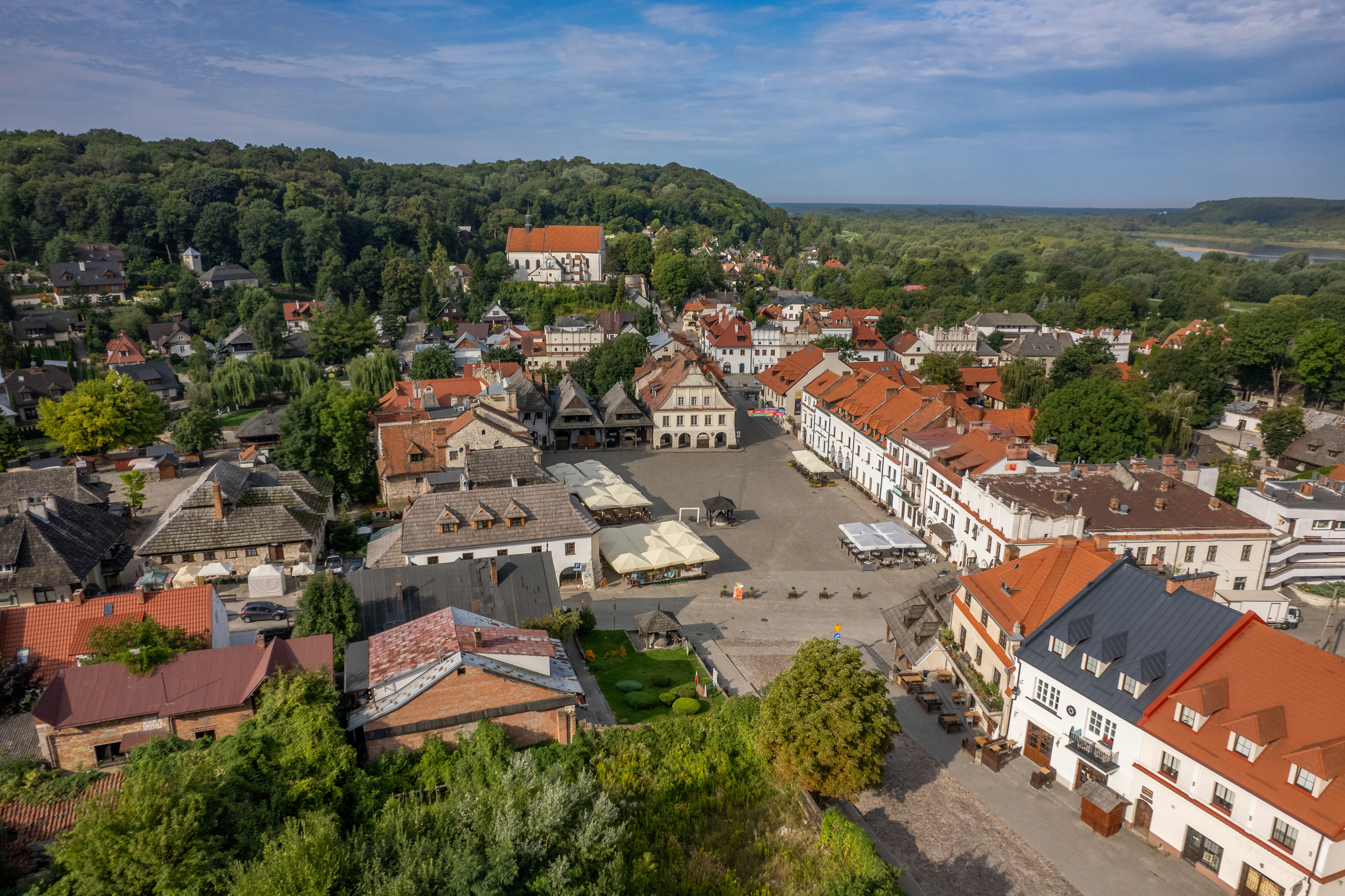 Widok z lotu ptaka na miasteczko z ryneczkiem otoczonym kolorowymi kamienicami. Na horyzoncie zalesione wzgórza i rzeka. Centrum zajmuje brukowany plac z namiotami kawiarnianymi. W tle widoczny jest duży budynek.