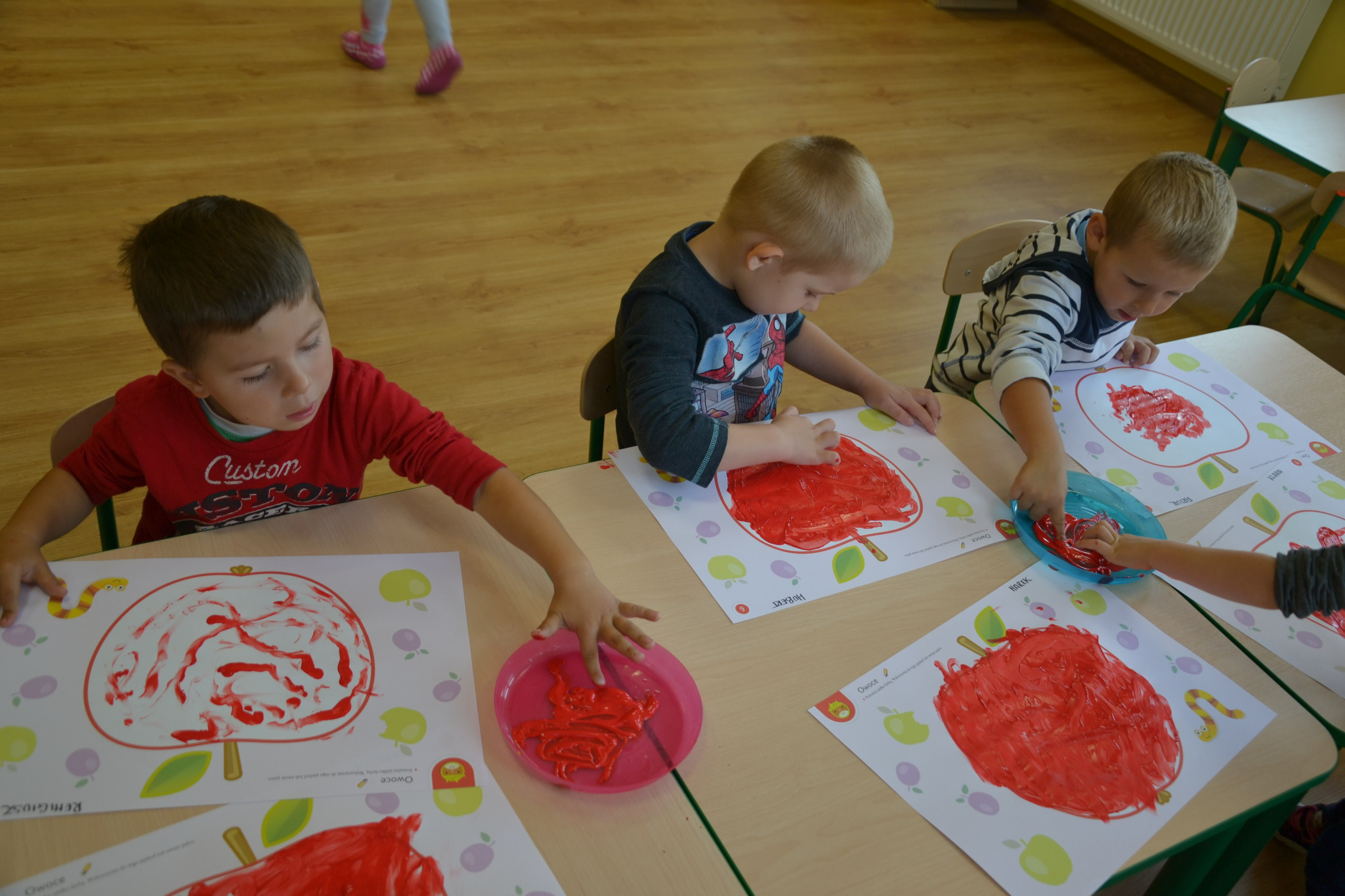 Trójka dzieci siedzi przy stole w przedszkolu, tworząc czerwone malunki palcami na kartkach. W pokoju widać zabawki i inne dzieci.