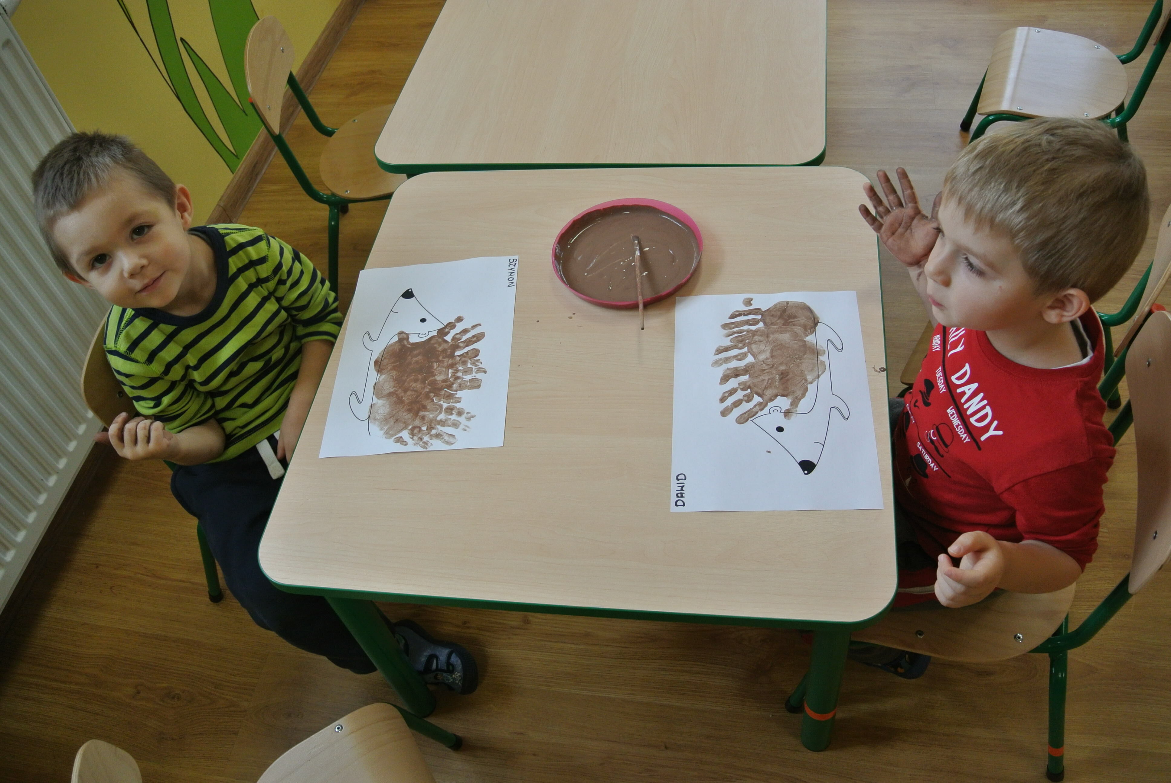 Dwóch chłopców siedzi przy stole z arkuszami papieru z odbitymi odciskami dłoni tworzącymi kształt zwierząt, jeden dziecko unosi rękę w geście pozdrowienia.