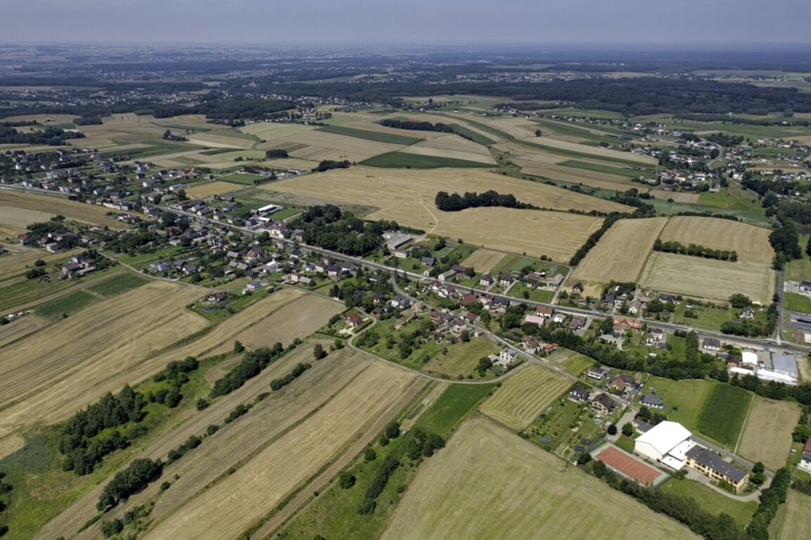 Zdjęcie lotnicze przedstawia wiejski krajobraz z polami, drogami i budynkami, zielonymi drzewami oraz rozległymi terenami uprawnymi.