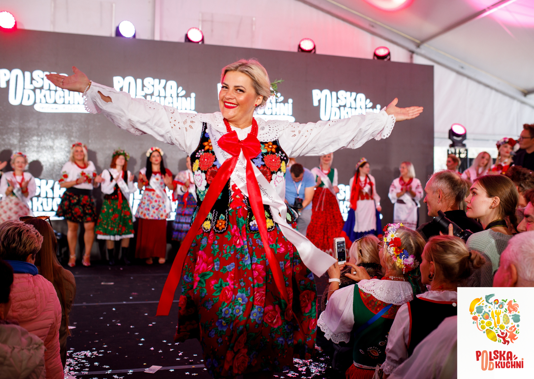 Kobieta w tradycyjnym, kolorowym polskim stroju ludowym z szeroko rozłożonymi ramionami tańczy, uśmiechając się. W tle tłum ludzi i inni tancerze.