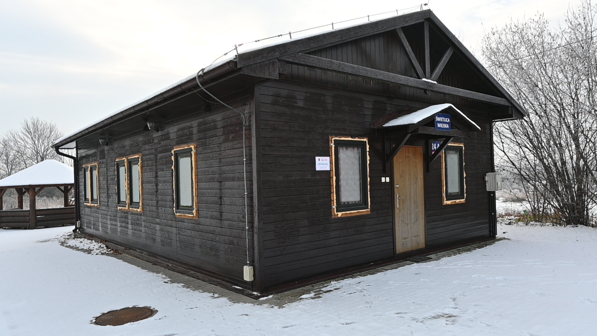 Drewniany budynek stacji kolejowej pokryty śniegiem, z oznakowaniem "Biletomat" i "WC", w zimowym krajobrazie.