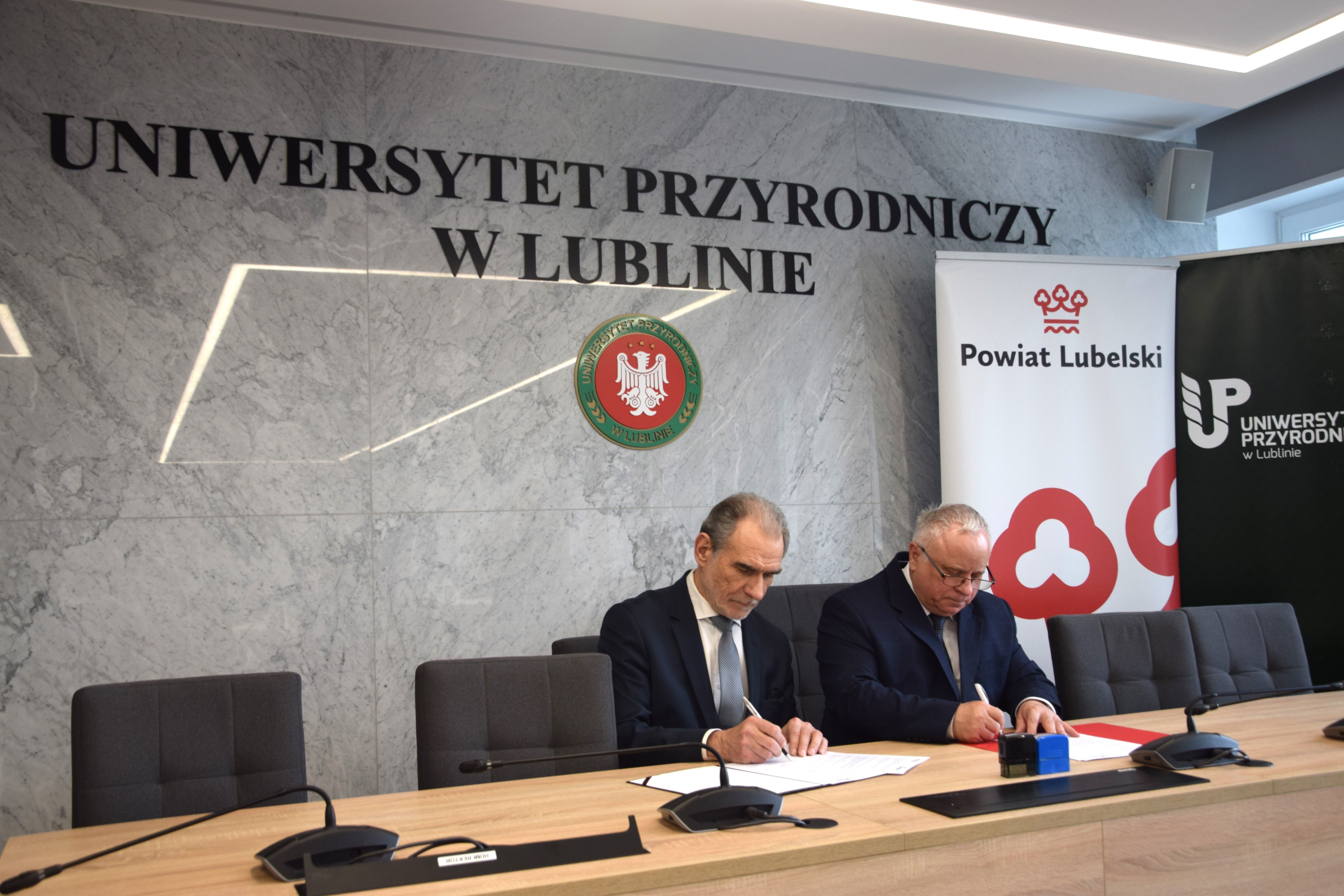 Dwóch mężczyzn podpisuje dokumenty w sali konferencyjnej z logotypami Uniwersytetu Przyrodniczego w Lublinie i Powiatu Lubelskiego.