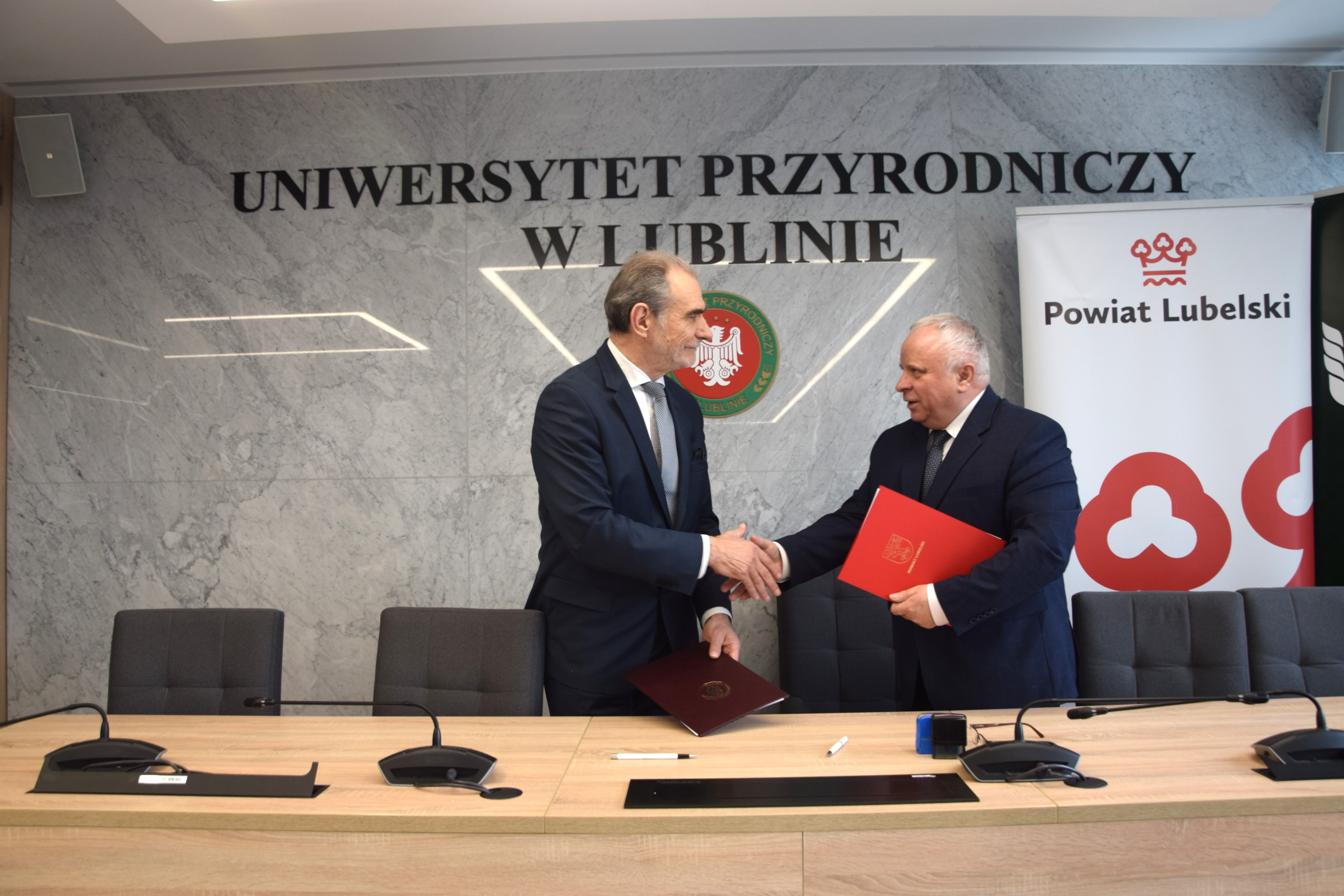 Dwóch mężczyzn w garniturach uściskuje sobie dłonie w sali konferencyjnej, po obu stronach stołu z mikrofonami, za nimi ściana z logo Uniwersytetu Przyrodniczego w Lublinie i Powiatu Lubelskiego.