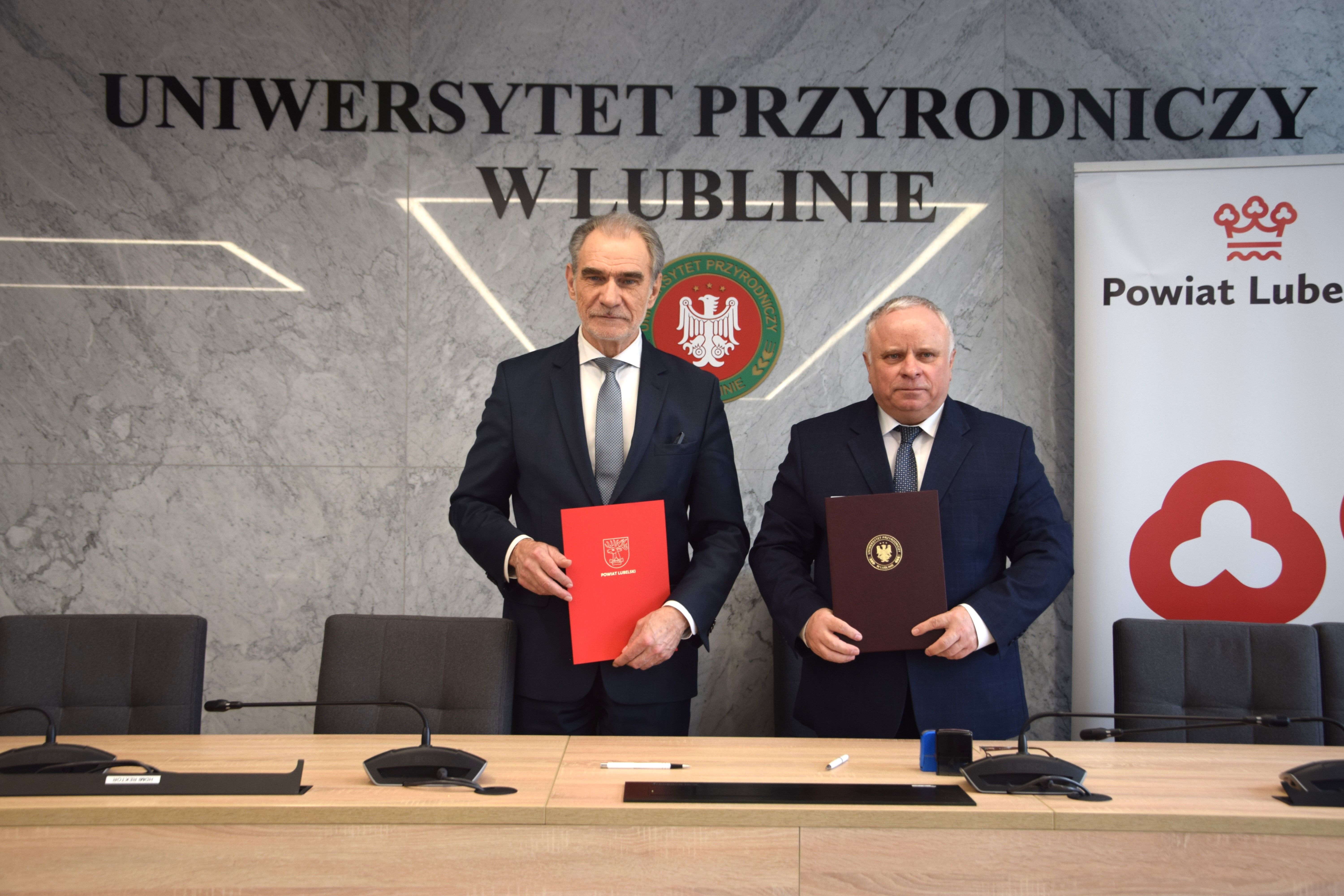 Dwaj mężczyźni ubrani w garnitury stoją w sali konferencyjnej, trzymają foldery, za nimi logo Uniwersytetu Przyrodniczego w Lublinie i herb Lublina.