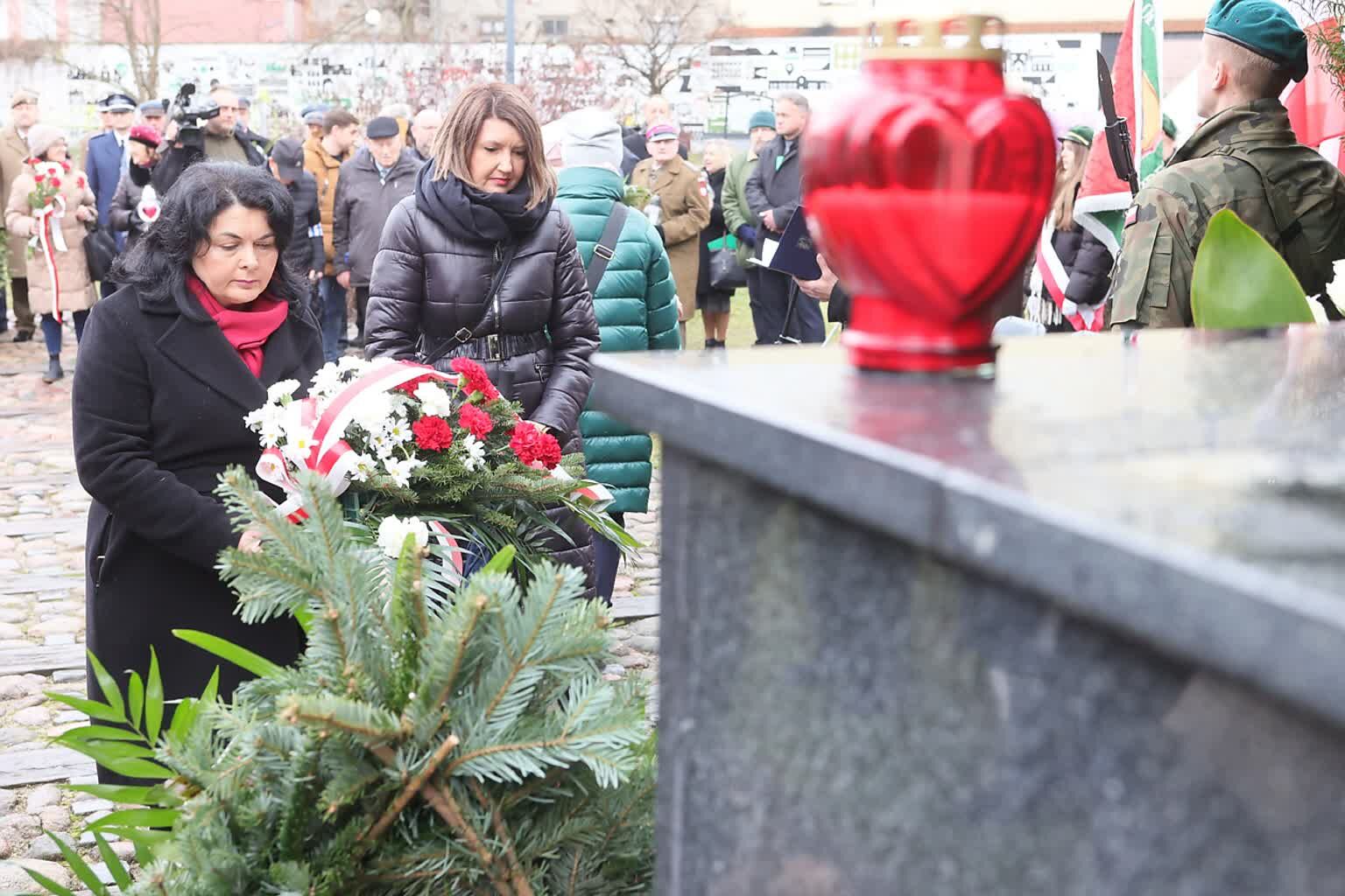 Na zdjęciu dwie kobiety składają kwiaty przy pomniku, w tle obecni są wojskowi, mamy widok na czerwoną znicz-lampę i gałęzie.