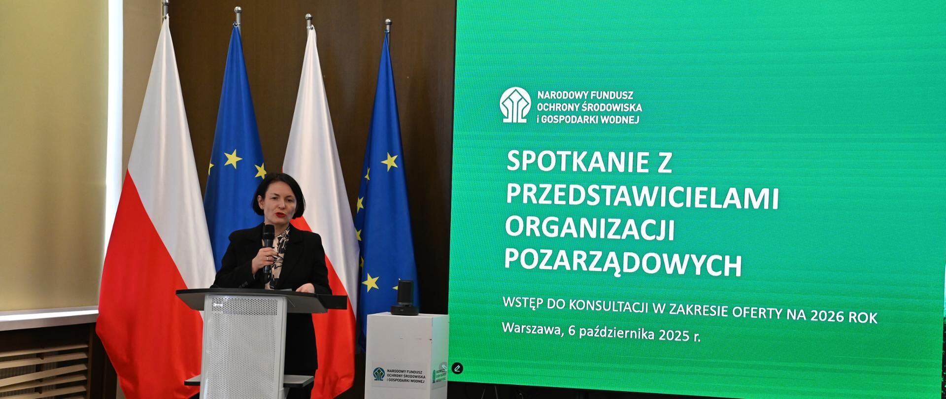 Na zdjęciu wystąpienie kobiety przy mównicy na tle polskiej, unijnej i innej flagi. Obok ekran z napisem o spotkaniu z organizacjami pozarządowymi dotyczącym konsultacji na 2026 rok. Data: 6 października 2023, Warszawa.