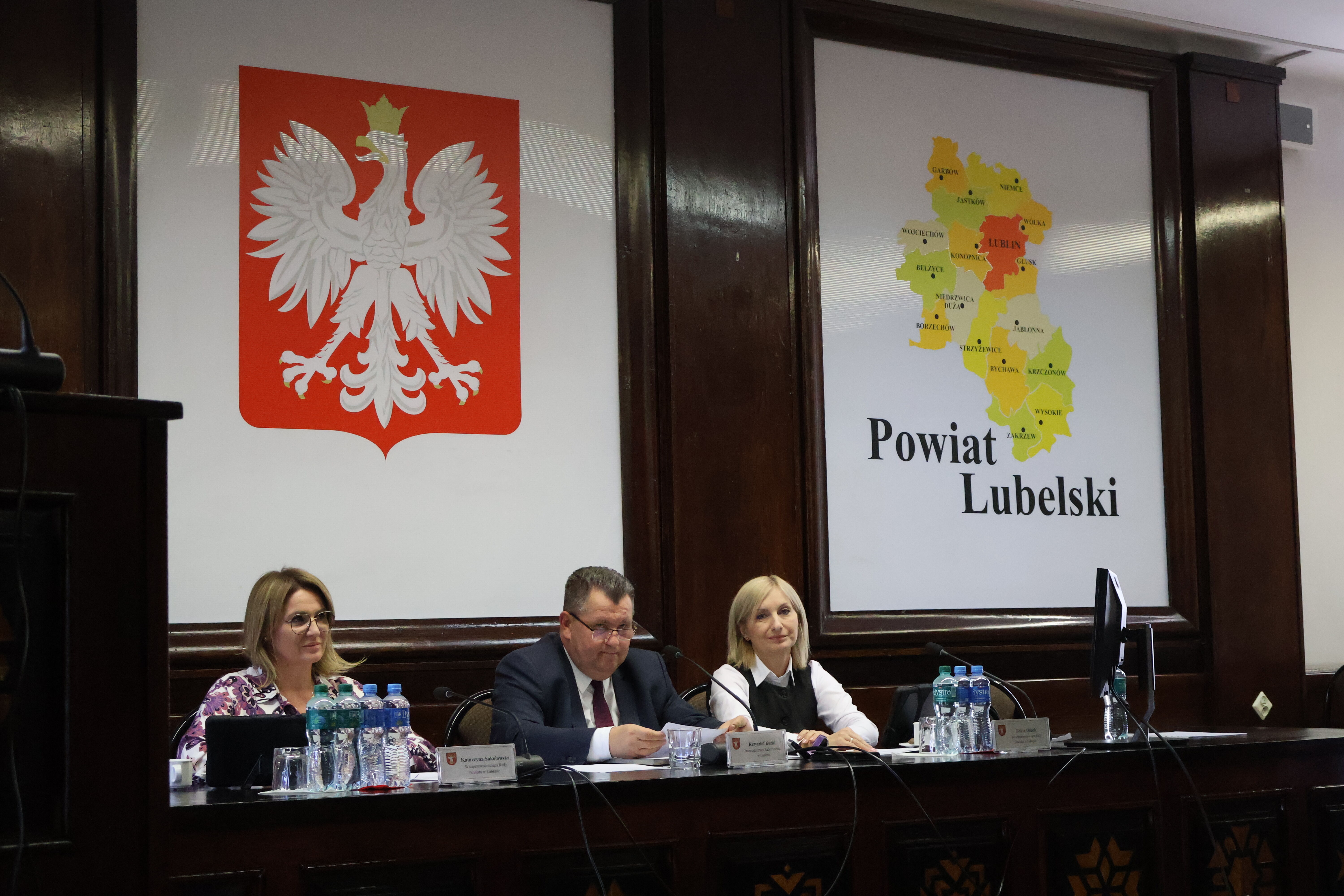 Trzy osoby siedzą za stołem w sali konferencyjnej. Na ścianie widoczne są godło Polski oraz mapa Powiatu Lubelskiego. Przed nimi stoją mikrofony i butelki z wodą.