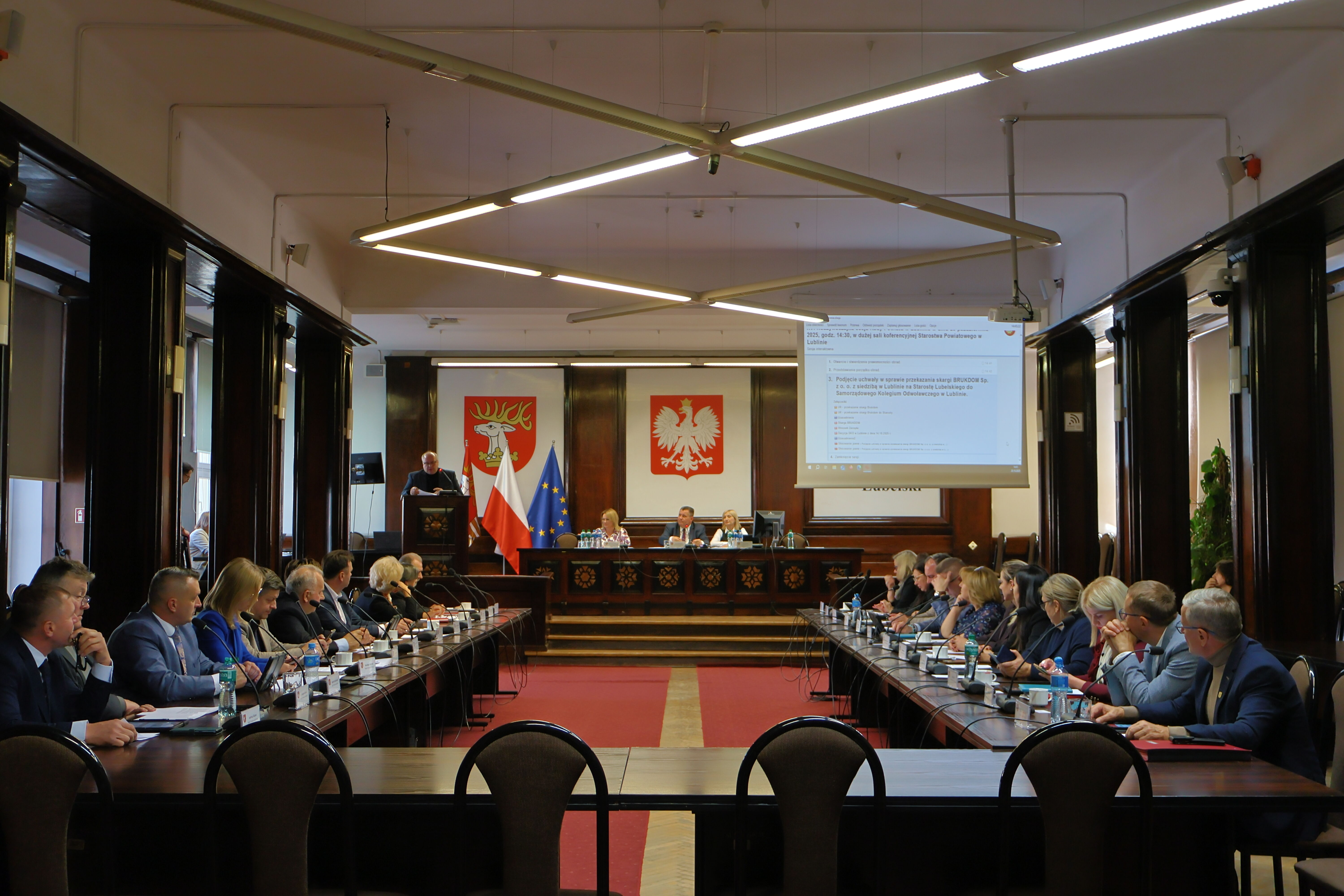 Sala obrad z długim stołem pełnym ludzi. Na końcu stołu widać mównicę i projektor wyświetlający tekst. Na ścianie flagi Polski i UE oraz godło Polski.