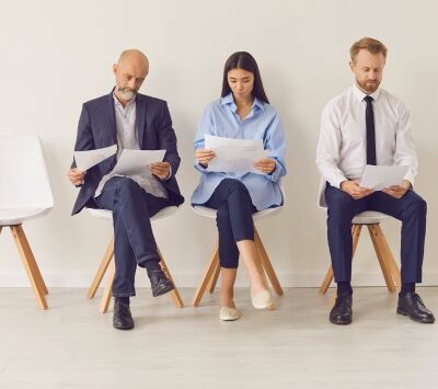 Trzy osoby siedzą na krzesłach w kolejce, przeglądając dokumenty. Wszyscy są elegancko ubrani, jak na rozmowę kwalifikacyjną. Tło jest białe, a podłoga drewniana.