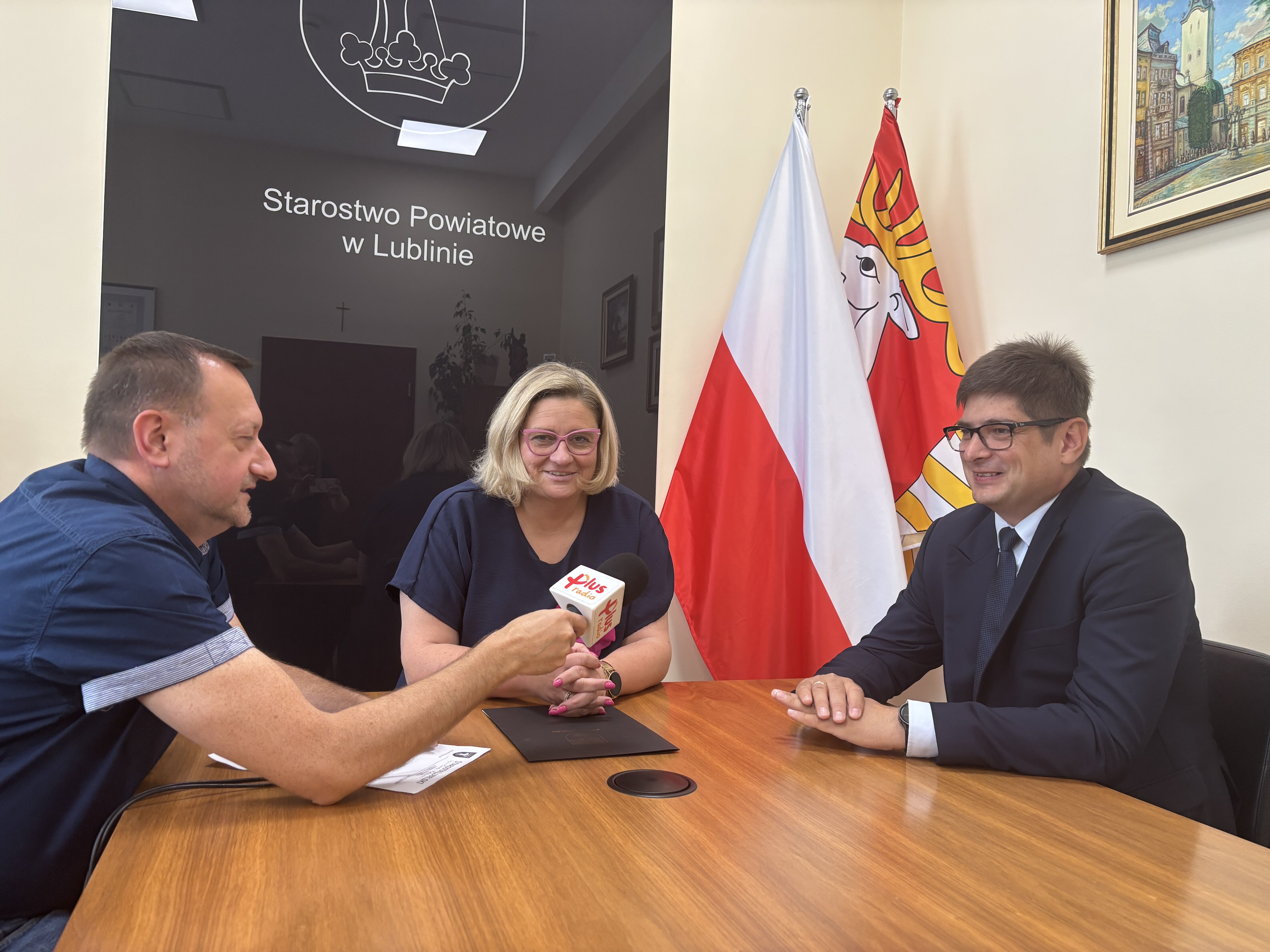 Na zdjęciu trzy osoby siedzą przy stole w biurze. Mężczyzna z mikrofonem rozmawia z kobietą i mężczyzną w garniturze. W tle widać flagi Polski i kolejnego regionu oraz napis "Starostwo Powiatowe w Lublinie".