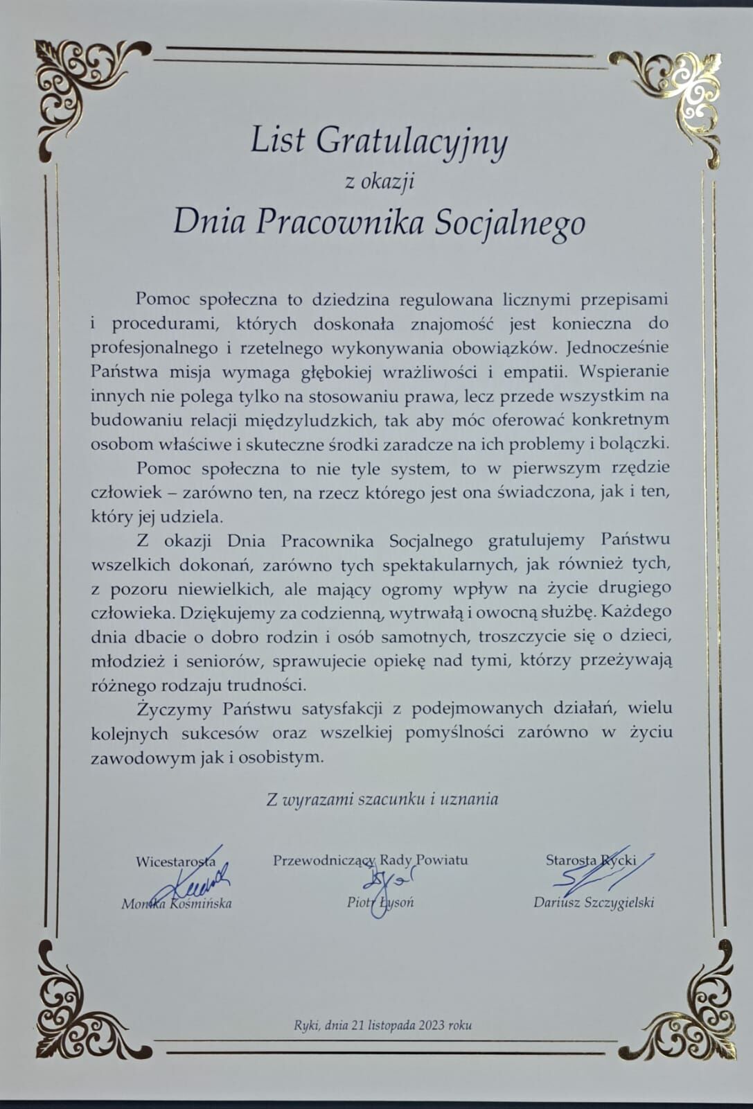 Zdjęcie przedstawia elegancki list gratulacyjny z ramką w ornamenty, wystawiony przez Dyrektora oraz Przewodniczącego Rady i Starostę Powiatu z okazji Dnia Pracownika Socjalnego.