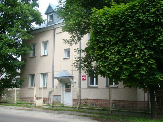Dom mieszkalny dwupiętrowy o jasnobrązowej fasadzie, z dachem dwuspadowym, otoczony drzewami, za metalowym ogrodzeniem, z numerem 22.