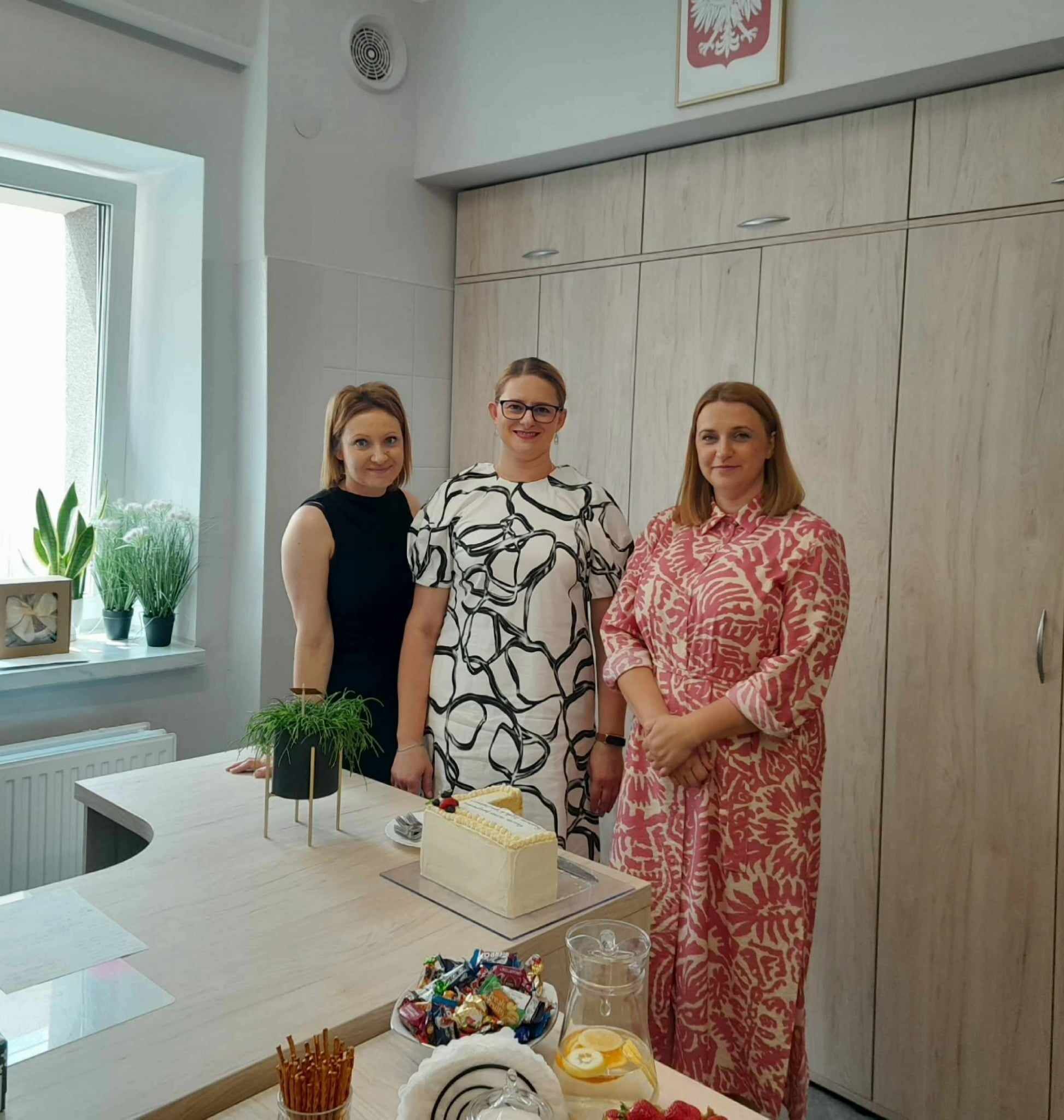 Trzy kobiety uśmiechają się do kamery, stojąc w jasnym pomieszczeniu, prawdopodobnie kuchni, z rośliną na blacie i polskim godłem na ścianie.