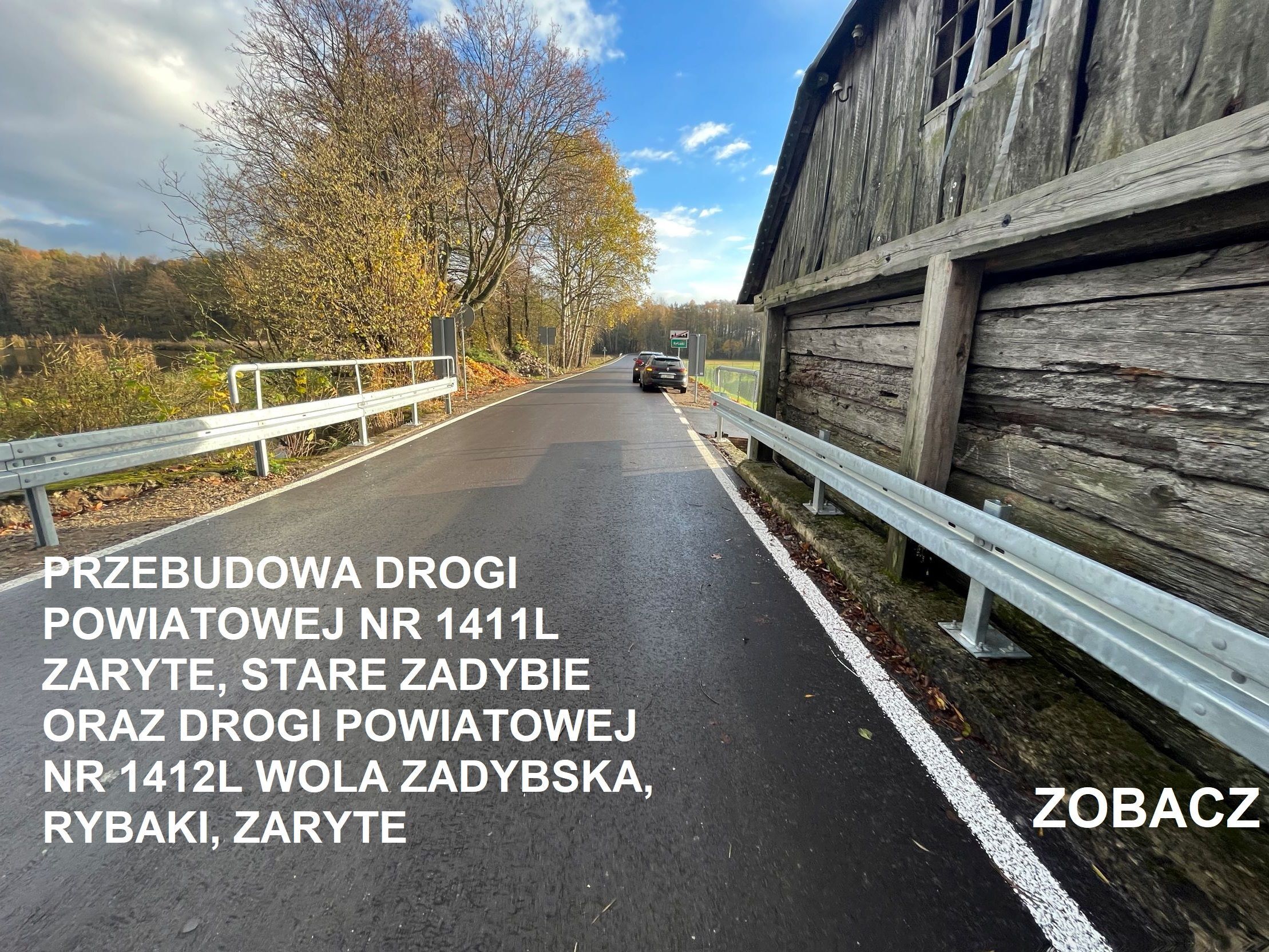 Droga asfaltowa z białą barierką obok drewnianej konstrukcji, z samochodami i lasem w tle, niebo z chmurami oraz informacyjny napis.