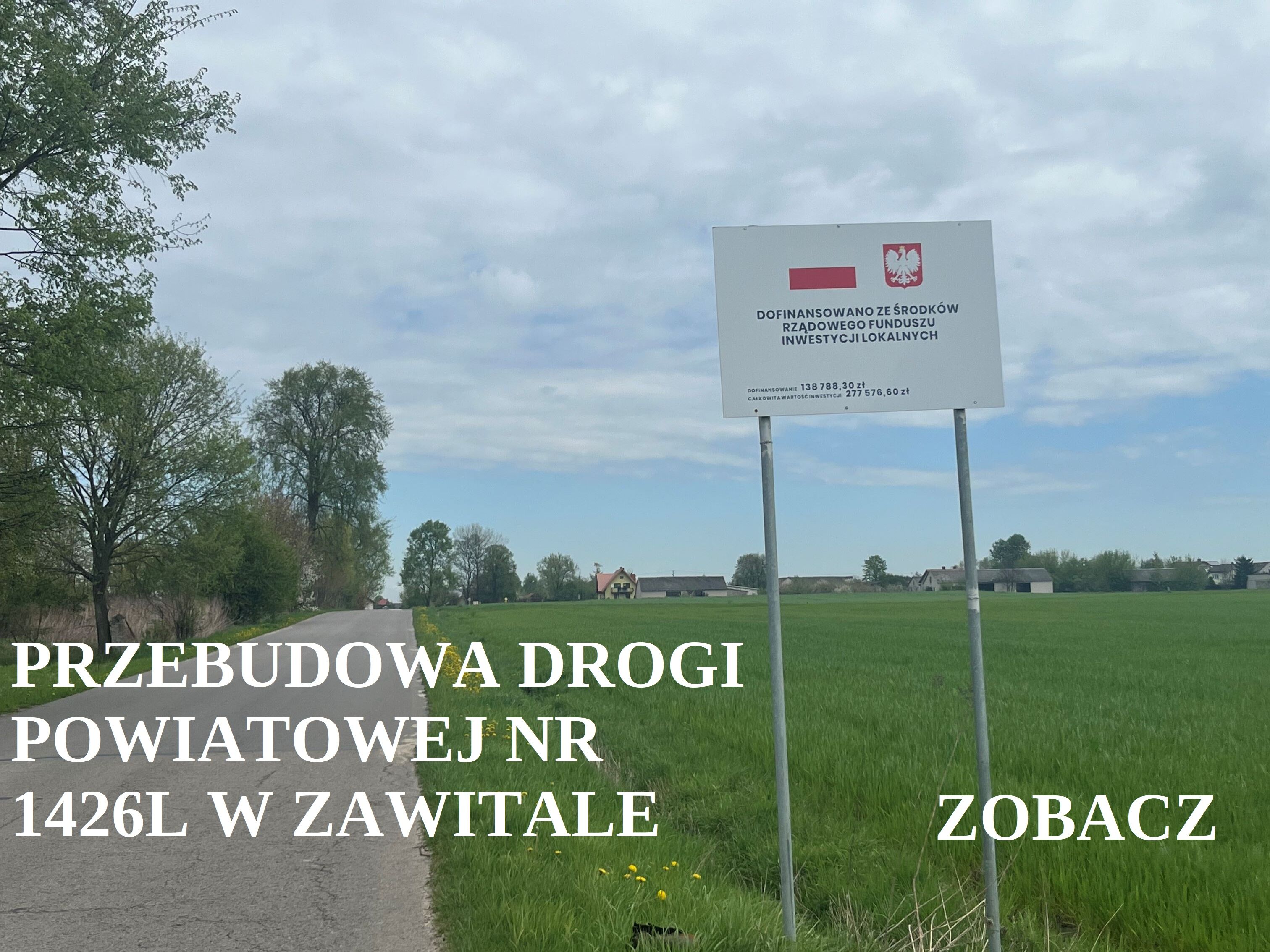 Tablica informacyjna przy drodze wiejskiej o przebudowie drogi powiatowej, zielone pola i rozproszone budynki w tle, niebo z chmurami.