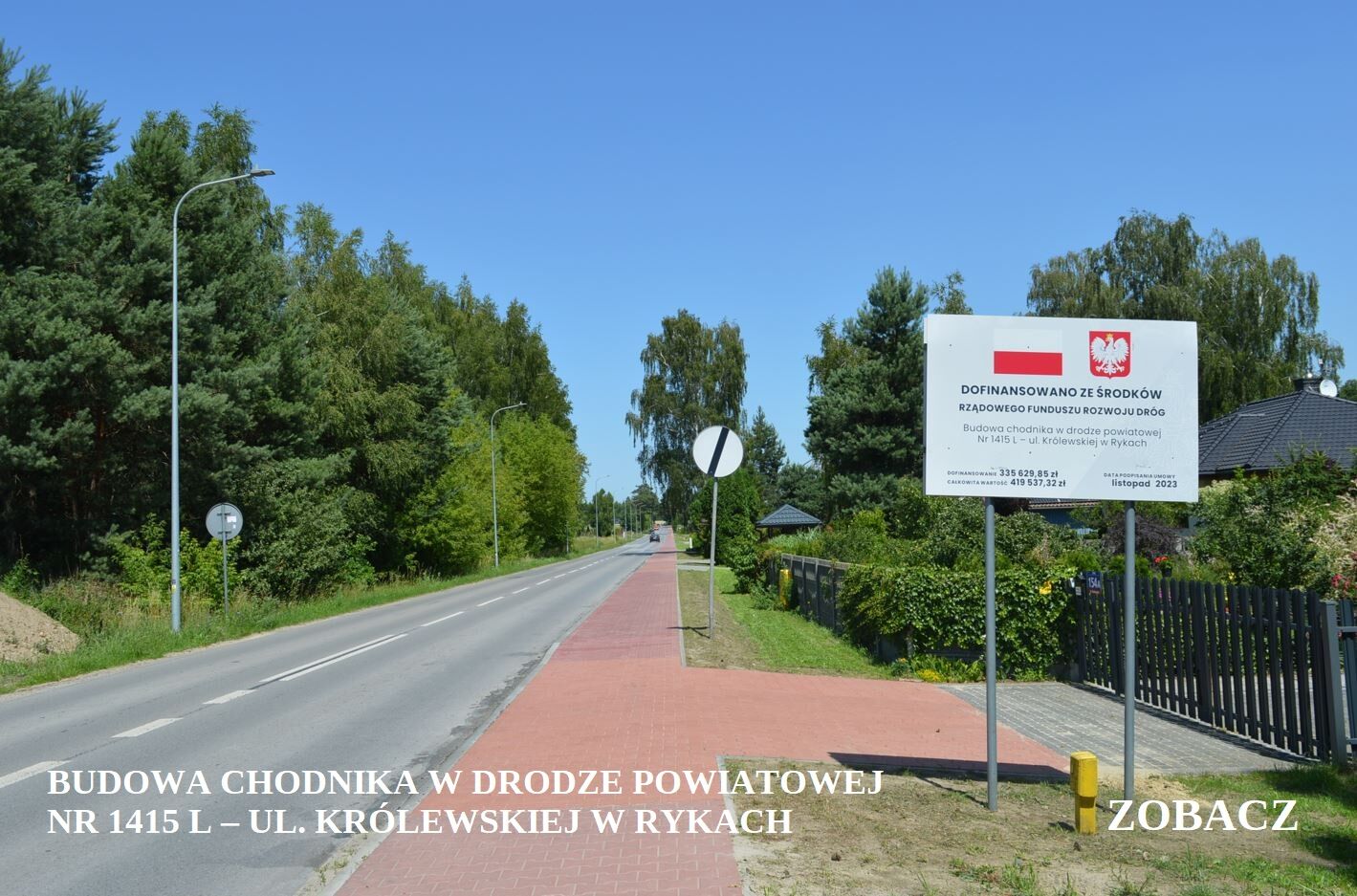 Droga asfaltowa z czerwonym chodnikiem biegnącym wzdłuż, otoczona zielenią z tablicą informacyjną po prawej stronie i znakiem drogowym.