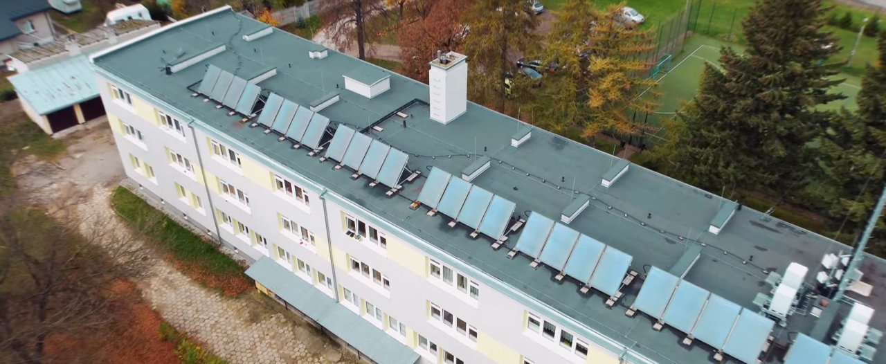Lotniczy widok szeregu paneli słonecznych na dachu budynku, z drzewami w tle.
