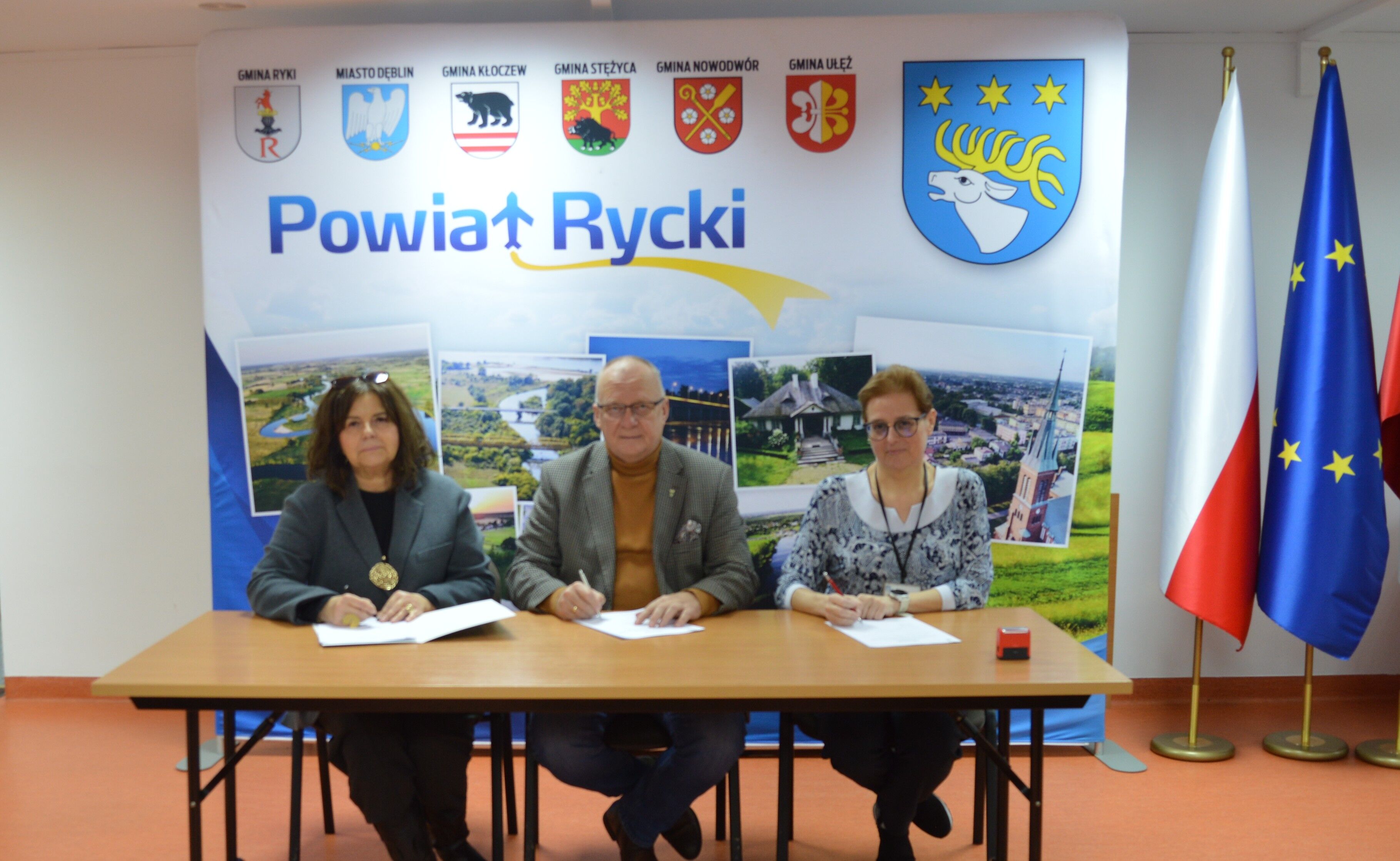 Na zdjęciu troje ludzi siedzi przy stole z dokumentami. W tle znajduje się baner z napisem „Powiat Rycki”, z herbami gmin i flagami Polski oraz Unii Europejskiej.