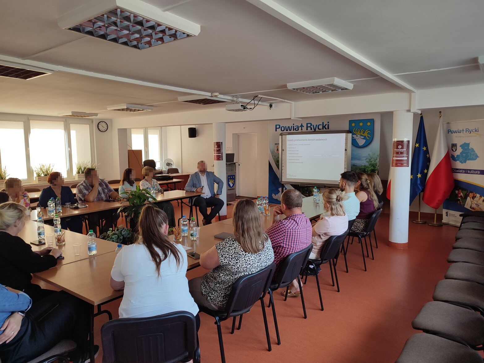 Spotkanie w sali konferencyjnej; ludzie siedzą wokół stołów, na których są butelki z wodą. Na ścianie widoczne logo Powiatu Ryckiego, obok polska flaga i ekran projekcyjny z prezentacją.