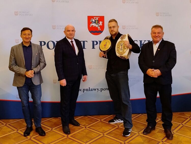 Spotkanie z mistrzem świata Jackiem Błasińskim w Starostwie Powiatowym w Puławach. Ludzie na tle ścianki z herbem Powiatu Puławskiego.