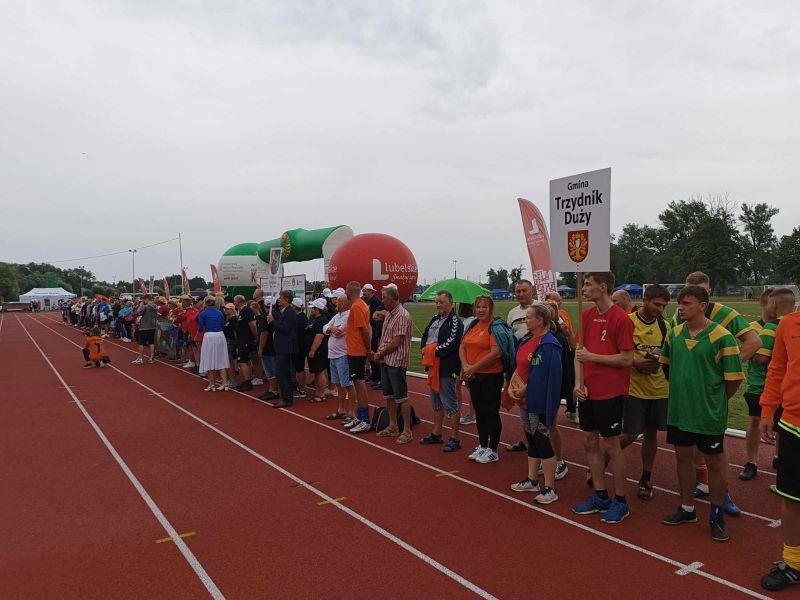 Uczestnicy wydarzenia stoją w kolejce na stadionie lekkoatletycznym, w tle namioty i banery reklamowe.