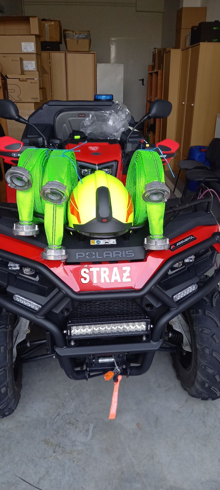 Czerwony quad z napisem "STRAŻ" na przedzie, wyposażony w zielony sprzęt ratowniczy, w tym kask. Pojazd stoi w garażu pełnym sprzętu.