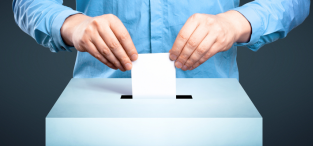 Osoba w niebieskiej koszuli wrzuca kartkę do głosowania do metalowej skrzynki na szarym tle, symbolizując oddanie głosu w wyborach.