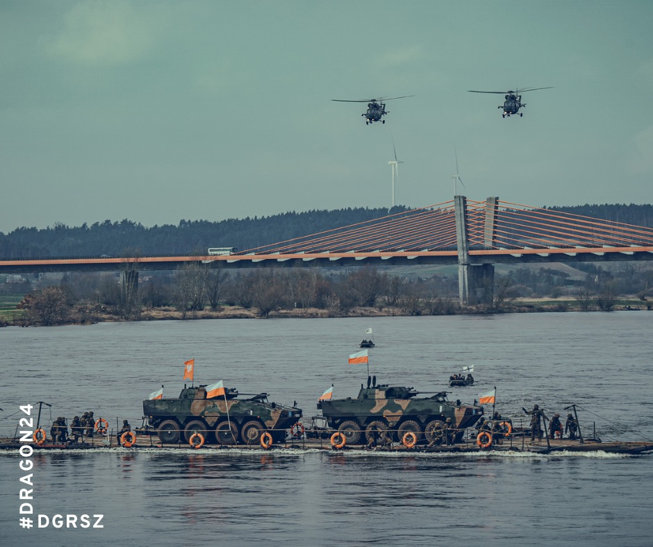 Zdjęcie przedstawia wojskowe pojazdy pancerne przemieszczające się na barkach przez rzekę, z dwoma helikopterami w powietrzu na tle mostu wiszącego.