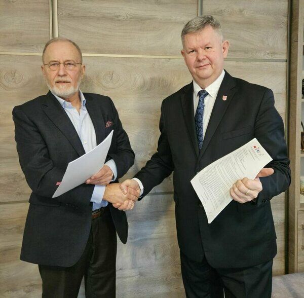 Dwóch mężczyzn stoi i uścisną się dłoni, trzymając dokumenty. Jeden w ciemnym garniturze, drugi w marynarce. Tło drewniana ściana.