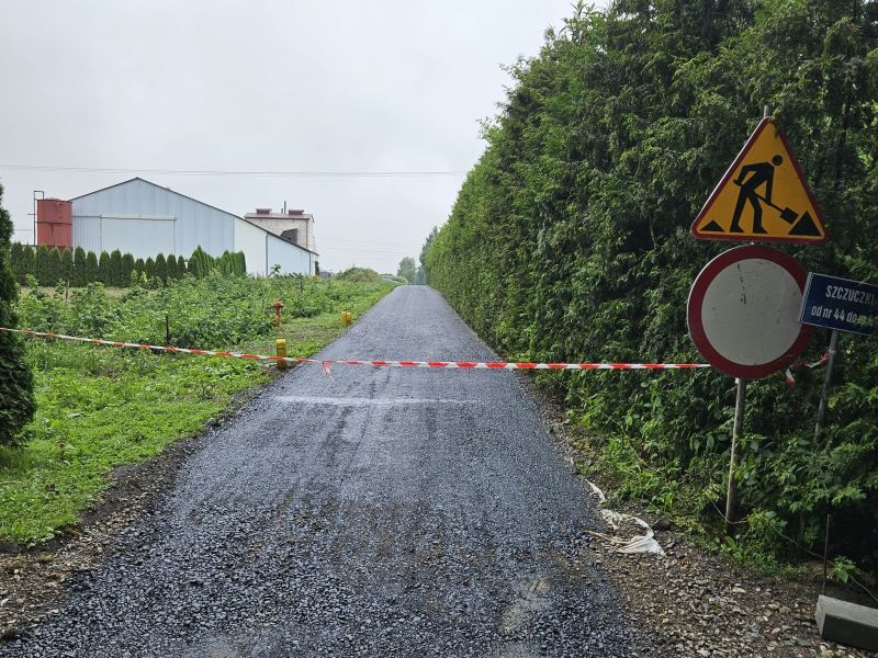 Droga z asfaltu zablokowana czerwono-białą taśmą, obok znaki drogowe: roboty i zakaz wjazdu. Zieleń po obu stronach, budynek w tle.