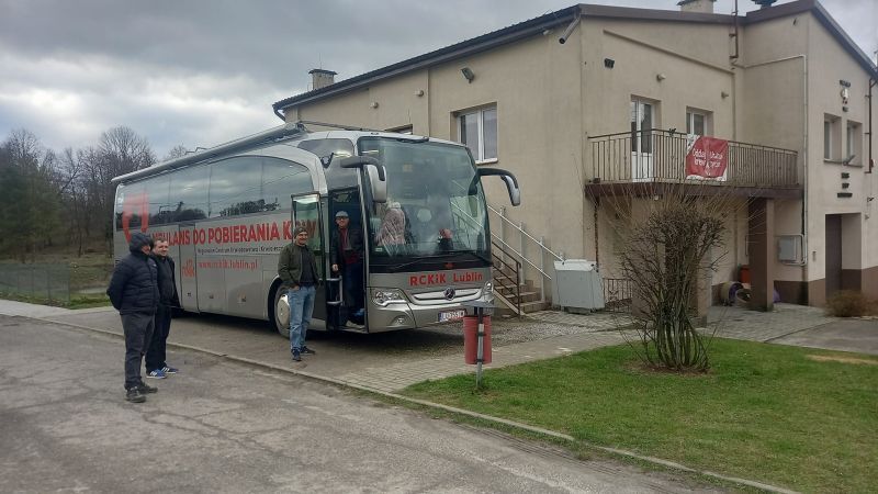 Autobus turystyczny zaparkowany przed budynkiem, z ludźmi wysiadającymi na parkingu obok drogi i trawnika. Chmurne niebo w tle.