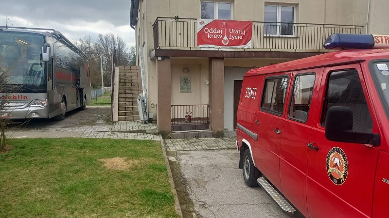 Autobus do poboru krwi zaparkowany obok budynku z plakatem zachęcającym do oddawania krwi, na pierwszym planie widoczny czerwony samochód straży pożarnej.